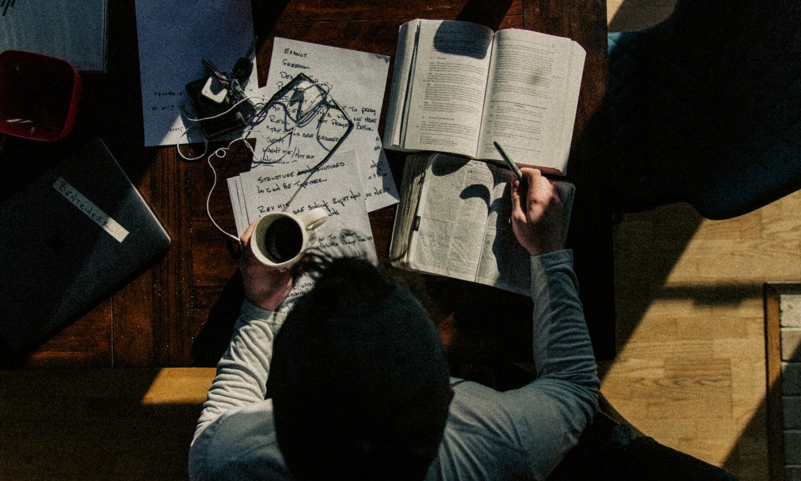 Homem visto de cima, estudando em uma escrivaninha com vários cadernos e papéis em cima, além de cabos de celular, uma xícara de café e um óculos