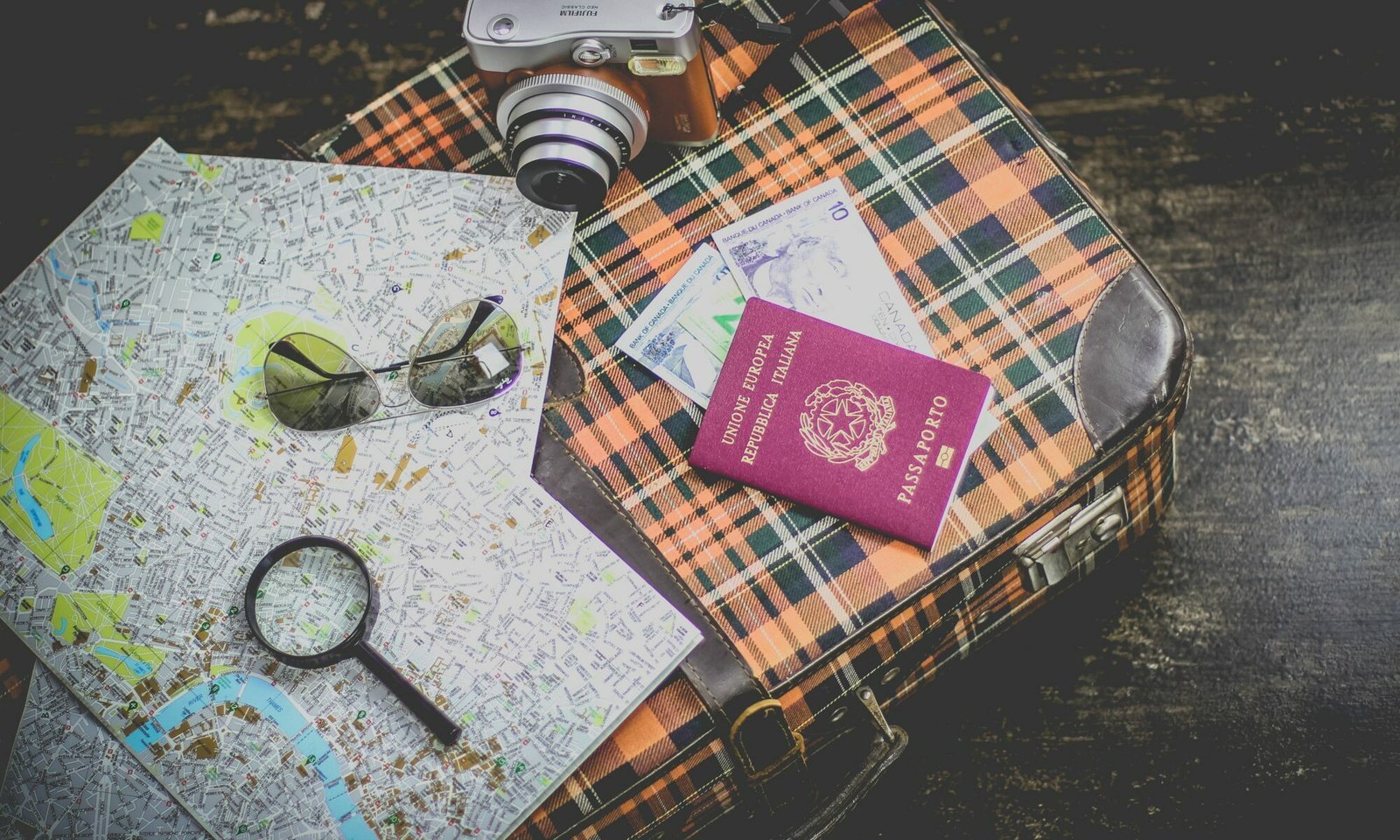 Mala de viagem com vários objetos em cima: um passaporte, uma lupa, mapas, um óculos de sol e uma câmera fotográfica