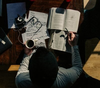 Homem visto de cima, estudando em uma escrivaninha com vários cadernos e papéis em cima, além de cabos de celular, uma xícara de café e um óculos